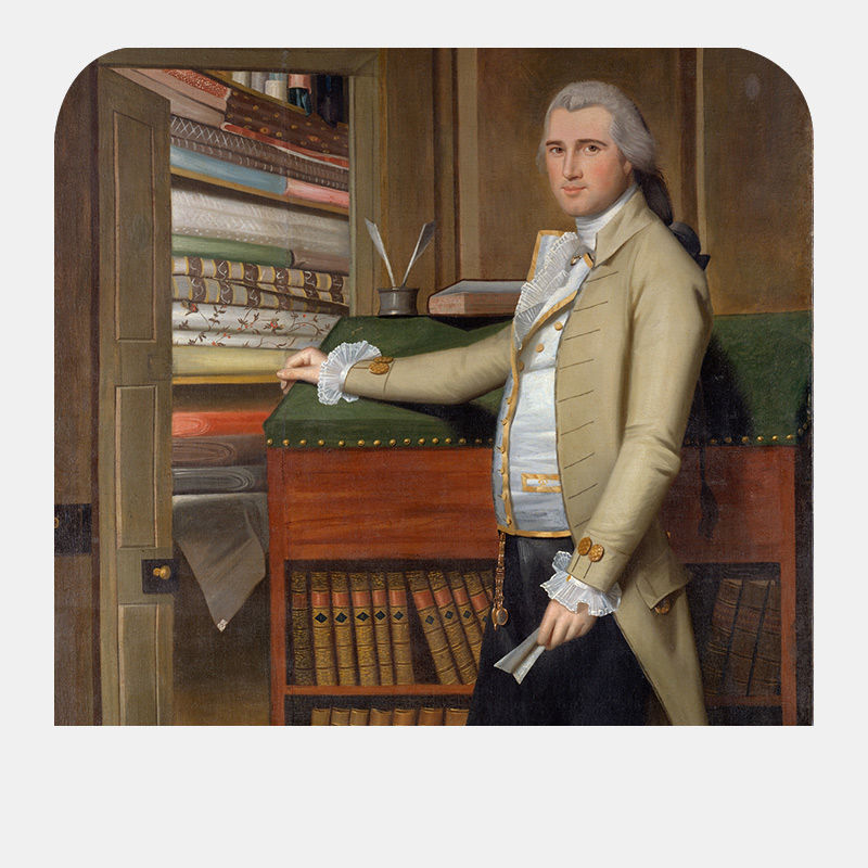 A man with gray hair pulled back and pale yellow coat stands next to an armoire of books, leaning his right arm on the surface as he holds a sheet of folded paper in his left hand.